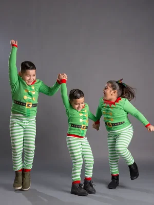 Three children in matching green and red elf pajamas holding hands and jumping playfully in unison against a grey background.