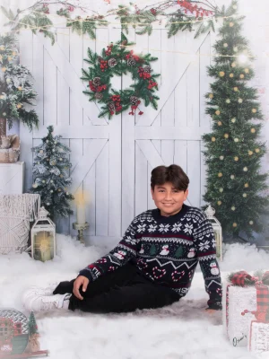 A boy sitting on the floor in a festive holiday sweater next to wrapped gifts and lanterns.