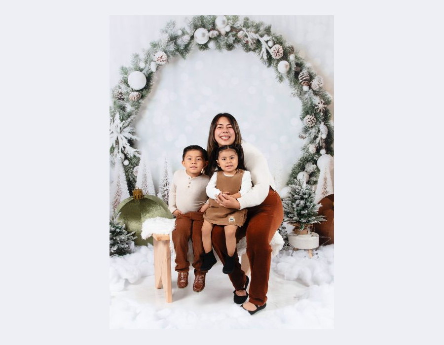 A woman and two children posing together on a snowy bench in front of a large frosted pine wreath arch.