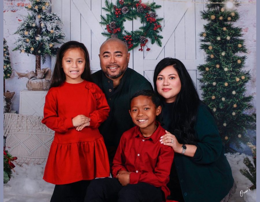 A family of four posing together in front of a white barn door backdrop adorned with a Christmas wreath and decorated trees.