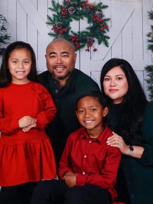 A family of four posing together in front of a white barn door backdrop adorned with a Christmas wreath and decorated trees.