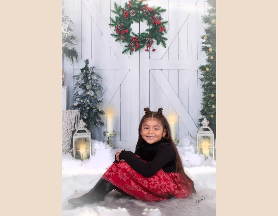 A young girl with hair buns sitting on the floor in a red and black outfit against a winter holiday backdrop with glowing lanterns and a wreath.