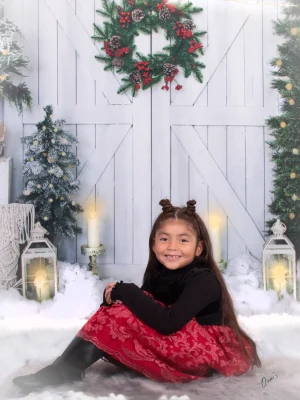 A young girl with hair buns sitting on the floor in a red and black outfit against a winter holiday backdrop with glowing lanterns and a wreath.