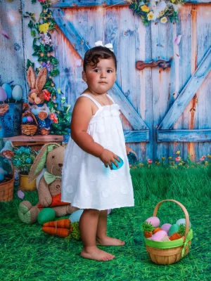 A young girl standing on green turf in front of a blue wooden door backdrop with Easter eggs, a basket, and a plush bunny.