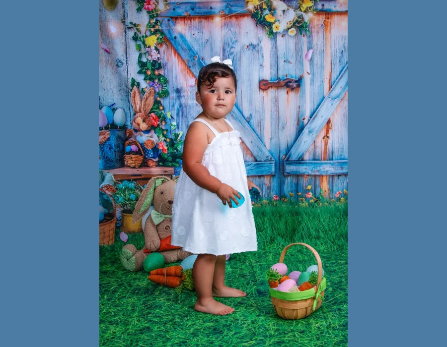 A young girl standing on green turf in front of a blue wooden door backdrop with Easter eggs, a basket, and a plush bunny.
