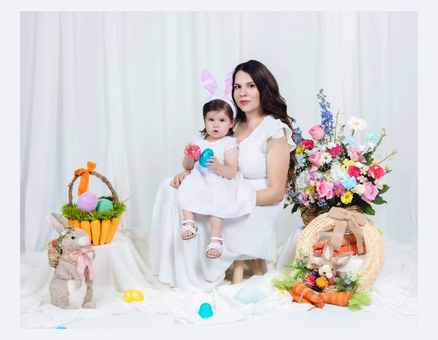 A woman holding a toddler wearing bunny ears in a studio set up with white curtains, a basket of eggs, floral arrangements, and a decorative rabbit.
