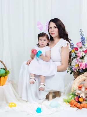 A woman holding a toddler wearing bunny ears in a studio set up with white curtains, a basket of eggs, floral arrangements, and a decorative rabbit.