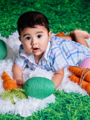 A baby lying on a pink rug with knit carrots and purple Easter eggs against a spring floral and grass backdrop.