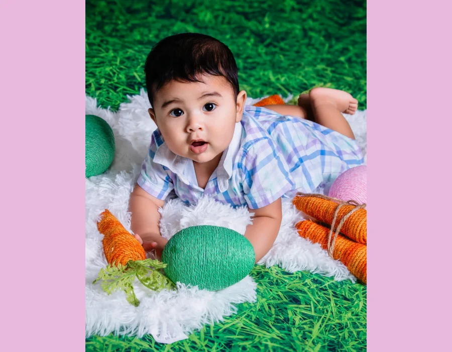 A baby lying on a pink rug with knit carrots and purple Easter eggs against a spring floral and grass backdrop.