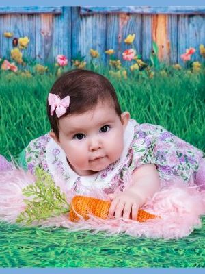 A baby lying on a white and green grass backdrop surrounded by yarn Easter eggs and knit carrots.