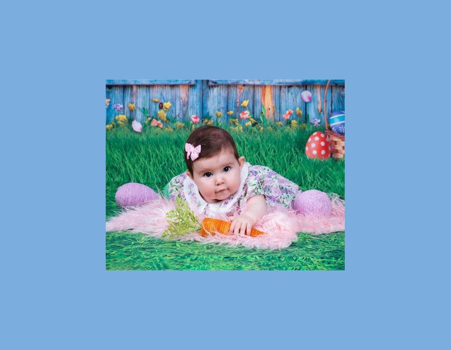 A baby lying on a white and green grass backdrop surrounded by yarn Easter eggs and knit carrots.