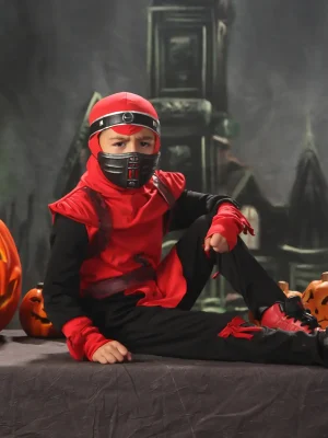 A young boy in a red and black ninja costume standing with his hands on his hips and fists clenched in front of a dark, spooky castle backdrop with pumpkins.