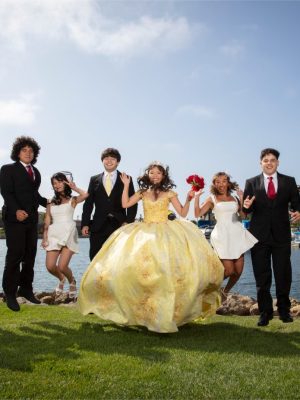 A large group of young adults in formal wear—including men in black suits and ties and women in short white dresses—along with a young woman in a large yellow ballgown, jumping in unison on a grassy lawn near a body of water.