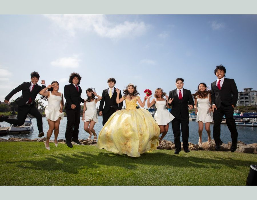 A large group of young adults in formal wear—including men in black suits and ties and women in short white dresses—along with a young woman in a large yellow ballgown, jumping in unison on a grassy lawn near a body of water.