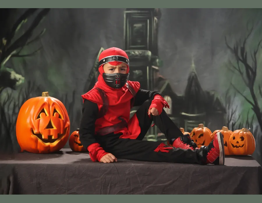 A young boy in a red and black ninja costume standing with his hands on his hips and fists clenched in front of a dark, spooky castle backdrop with pumpkins.