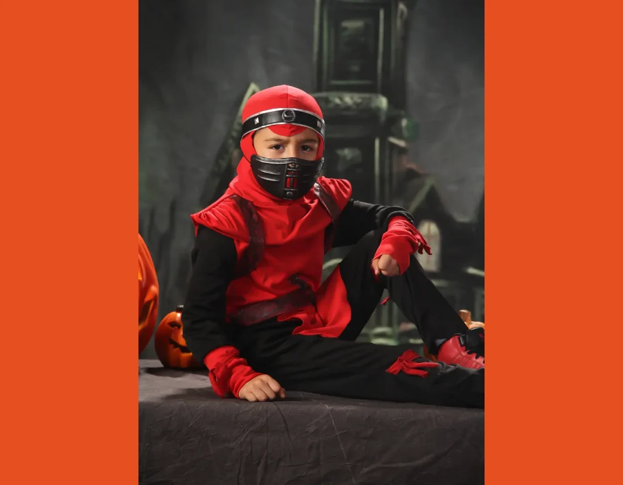 A young boy wearing a red and black ninja costume and mask, sitting on a dark draped table next to several carved jack-o'-lantern pumpkins against a spooky castle backdrop.