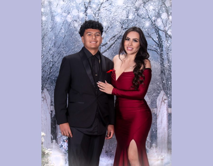 : A young man in a black tuxedo and a woman in a glamorous off-the-shoulder red dress with a thigh-high slit posing together in front of a snowy winter backdrop.
