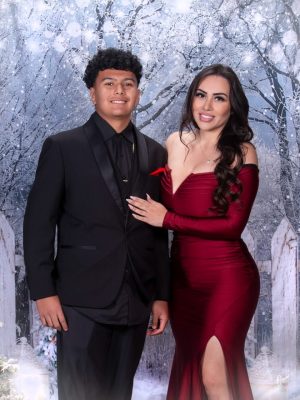 : A young man in a black tuxedo and a woman in a glamorous off-the-shoulder red dress with a thigh-high slit posing together in front of a snowy winter backdrop.