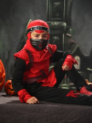 A young boy wearing a red and black ninja costume and mask, sitting on a dark draped table next to several carved jack-o'-lantern pumpkins against a spooky castle backdrop.