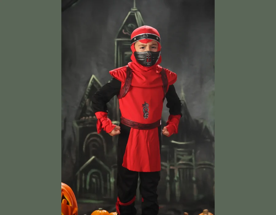 A close-up portrait of a young boy in a red and black ninja costume with his hands in fists, sitting on a dark table next to a jack-o'-lantern against a spooky background.