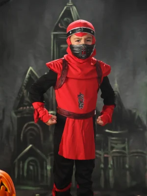 A close-up portrait of a young boy in a red and black ninja costume with his hands in fists, sitting on a dark table next to a jack-o'-lantern against a spooky background.