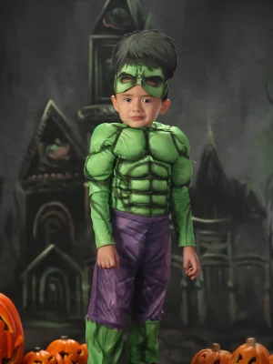 A young boy dressed in a Hulk costume complete with a muscle-padded top, purple pants, and a green mask, standing in front of a dark, spooky castle backdrop with lit jack-o'-lanterns.