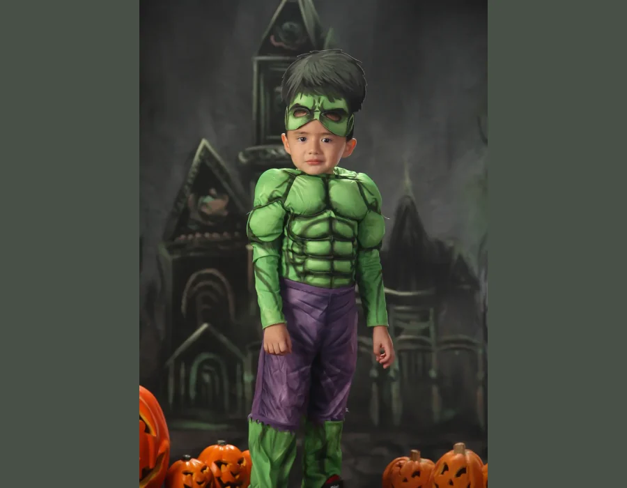 A young boy dressed in a Hulk costume complete with a muscle-padded top, purple pants, and a green mask, standing in front of a dark, spooky castle backdrop with lit jack-o'-lanterns.