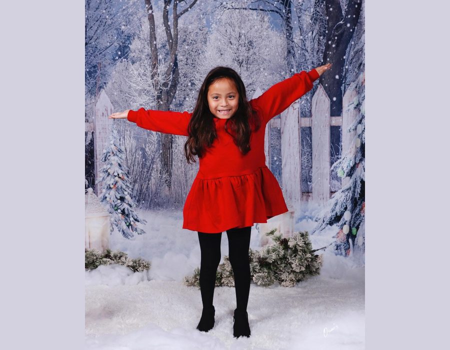 child posing with arms outstretched against a snowy winter-themed studio backdrop with a white picket fence and Christmas trees