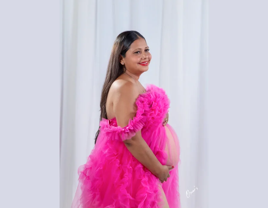A pregnant woman wearing an off-the-shoulder vibrant pink ruffled gown, smiling as she cradles her baby bump against a white backdrop.
