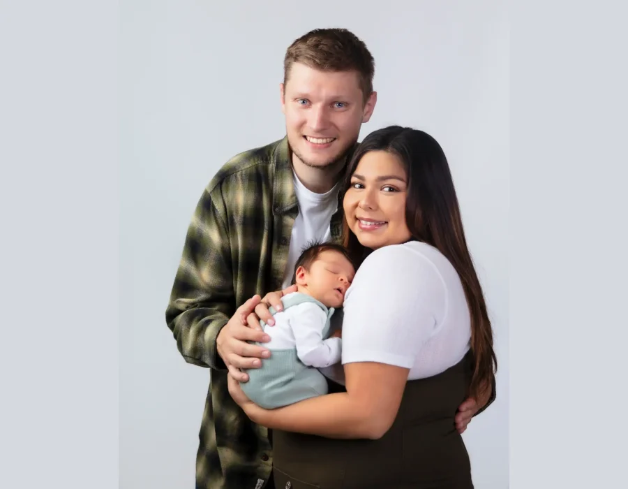 A happy family portrait in the studio, showing a mother and father smiling at the camera while holding their newborn baby who is sleeping peacefully in sage green overalls.