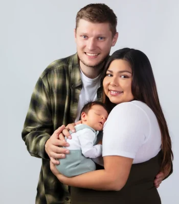 A happy family portrait in the studio, showing a mother and father smiling at the camera while holding their newborn baby who is sleeping peacefully in sage green overalls.