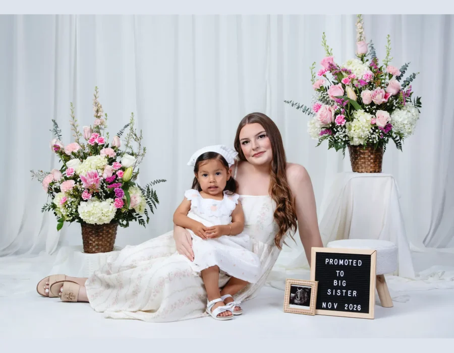 A pregnant woman sitting on a white studio floor in a white dress, holding a young girl in a matching white dress on her lap, next to a sign that reads "Promoted to Big Sister Nov 2026" and an ultrasound photo, surrounded by large floral arrangements.