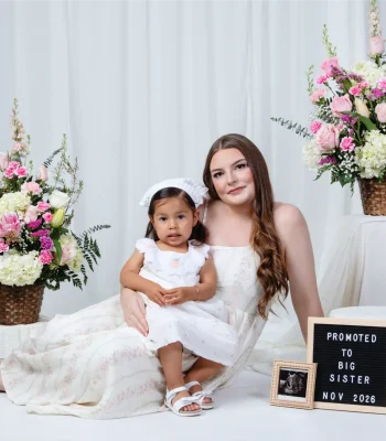 A pregnant woman sitting on a white studio floor in a white dress, holding a young girl in a matching white dress on her lap, next to a sign that reads "Promoted to Big Sister Nov 2026" and an ultrasound photo, surrounded by large floral arrangements.