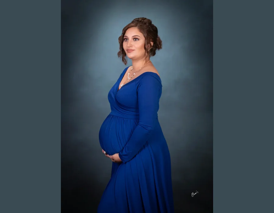A profile studio portrait of a pregnant woman wearing an elegant long-sleeve royal blue dress with her hair styled in an updo, gently supporting her baby bump against a dark blue textured background. WEBP