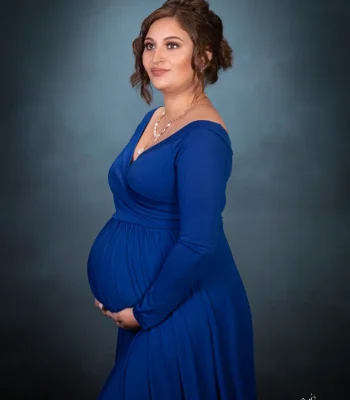 A profile studio portrait of a pregnant woman wearing an elegant long-sleeve royal blue dress with her hair styled in an updo, gently supporting her baby bump against a dark blue textured background. WEBP