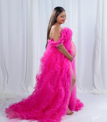 A full-length portrait of a smiling pregnant woman in a dramatic, long-trained off-the-shoulder pink ruffled dress posing sideways in a studio.