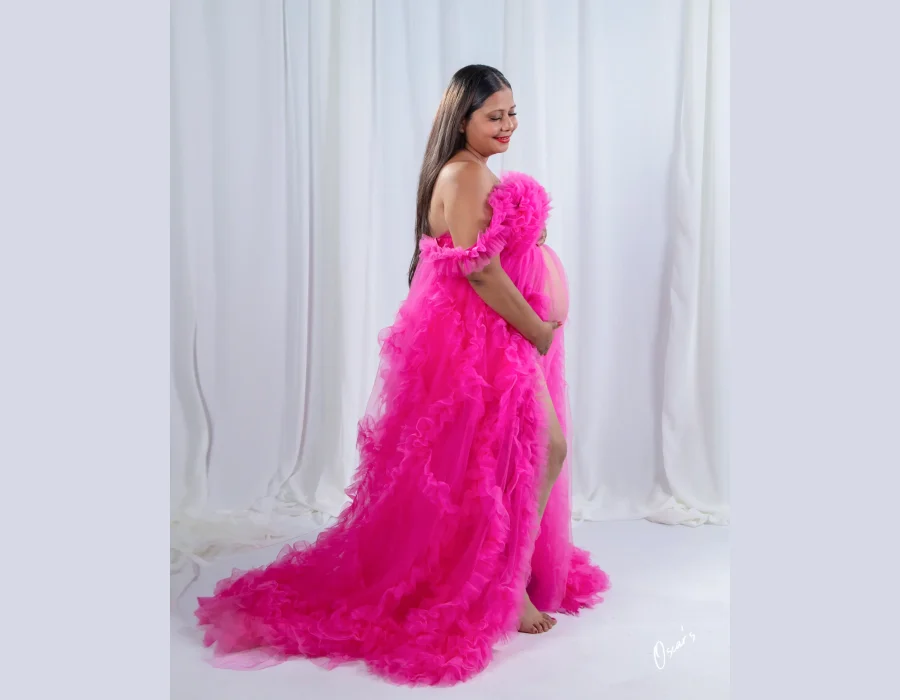 A full-length portrait of a smiling pregnant woman in a dramatic, long-trained off-the-shoulder pink ruffled dress posing sideways in a studio.