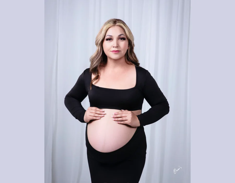 A portrait of a pregnant woman wearing a long-sleeve black crop top and skirt, gently holding her exposed belly with both hands.