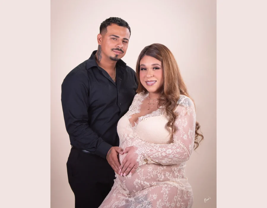 A couple posing together in a studio, with a man in a black shirt holding hands with a pregnant woman in a cream-colored lace dress to form a heart shape over her bump.