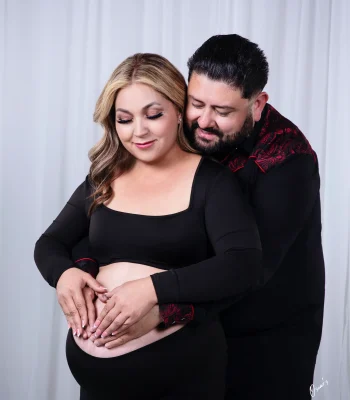 An expectant couple embracing in the studio, with the partner wrapping his arms around the pregnant woman in black and both placing their hands in a heart shape on her belly.