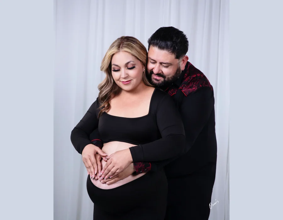 An expectant couple embracing in the studio, with the partner wrapping his arms around the pregnant woman in black and both placing their hands in a heart shape on her belly.