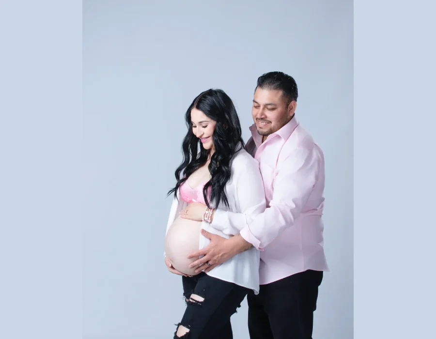 A smiling pregnant woman in a white button-down shirt and pink lace bra, with her partner standing behind her and placing his hands over her bump against a light background.