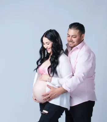 A smiling pregnant woman in a white button-down shirt and pink lace bra, with her partner standing behind her and placing his hands over her bump against a light background.