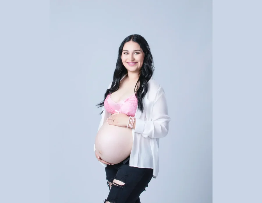 A close-up studio portrait of a pregnant woman with dark hair wearing an open white shirt over a pink lace bra, smiling down at her baby bump.