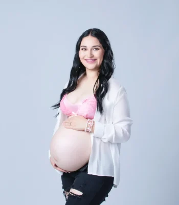 A close-up studio portrait of a pregnant woman with dark hair wearing an open white shirt over a pink lace bra, smiling down at her baby bump.