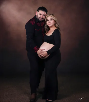 A couple posing for a studio maternity portrait against a textured brown backdrop, wearing matching black outfits while holding the pregnant woman's belly together.