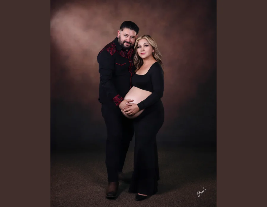 A couple posing for a studio maternity portrait against a textured brown backdrop, wearing matching black outfits while holding the pregnant woman's belly together.