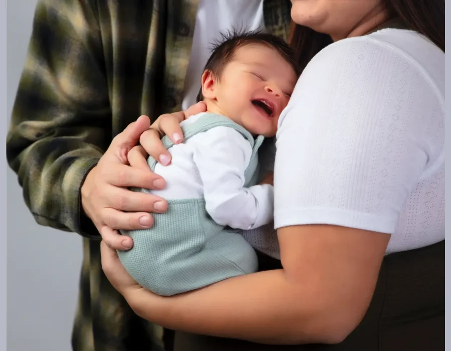 A close-up shot of a newborn baby in green overalls smiling softly while resting against the mother's shoulder, with the father's hands gently supporting the baby.