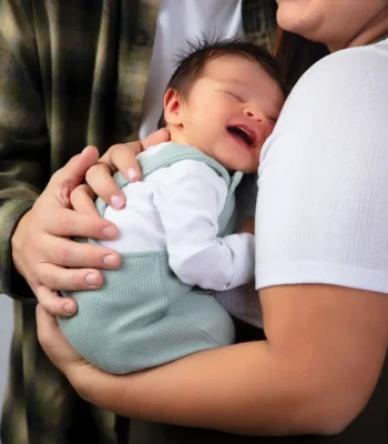 A close-up shot of a newborn baby in green overalls smiling softly while resting against the mother's shoulder, with the father's hands gently supporting the baby.