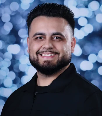 A close-up studio portrait of a smiling man with short dark hair, a beard, and a mustache, wearing a black shirt against a blue bokeh background.